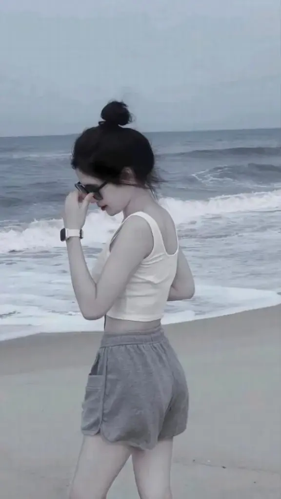 Ảnh cô gái đi biển giấu mặt, tay che nắng, váy xòe mềm, gió biển làm tóc rối.