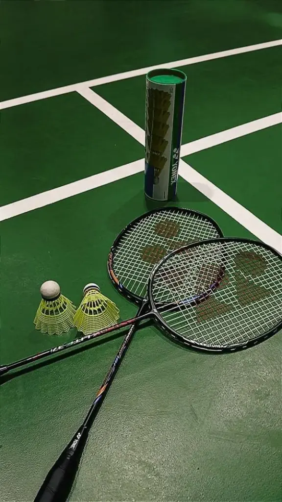 Hai chiếc vợt cầu lông nằm cạnh nhau trên mặt thảm sân xanh mướt dưới ánh đèn sân vận động tạo nên một không gian thi đấu chân thực nhất.