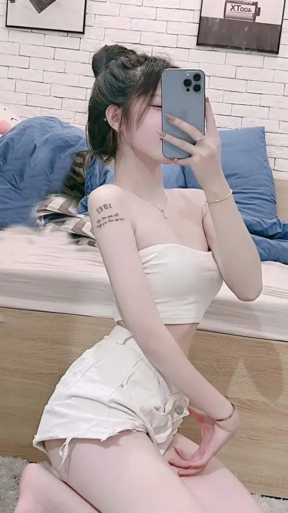 Nàng ngồi nghiêng trên giường, hông cong tinh tế, đùi trắng mịn, gương mặt che nhẹ vẫn quyến rũ